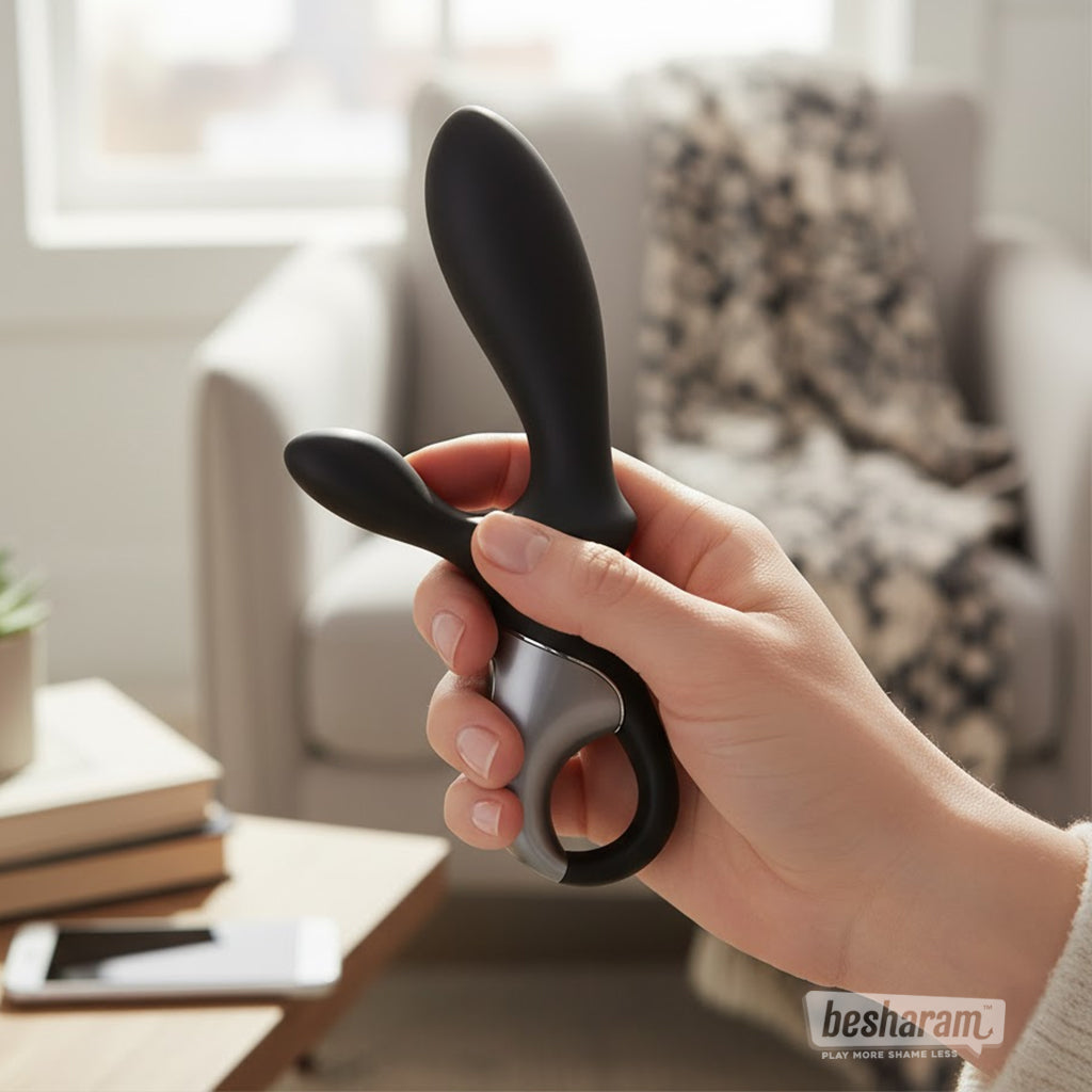 Hand holding the Satisfyer Heat Climax+ anal vibrator in a cozy living room setting, showing its sleek black dual-arm design.