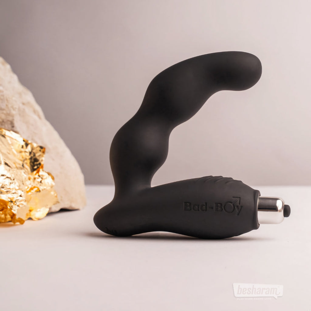 The black Bad-Boy prostate massager standing upright on a neutral backdrop with decorative stones and gold textures.