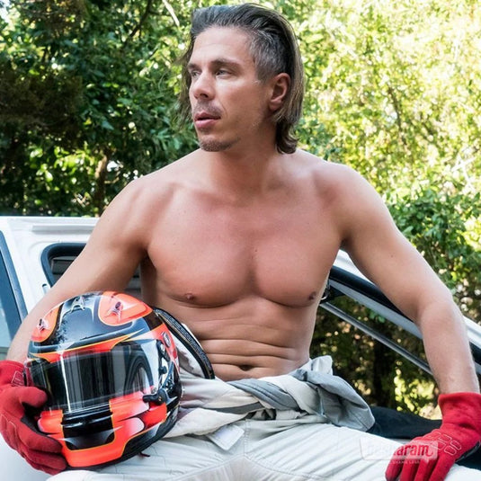 Mick Blue sitting shirtless beside a car holding a racing helmet in an outdoor setting