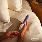 Hand holding the LELO LIV 3 Plum vibrator on a bed with white pillows