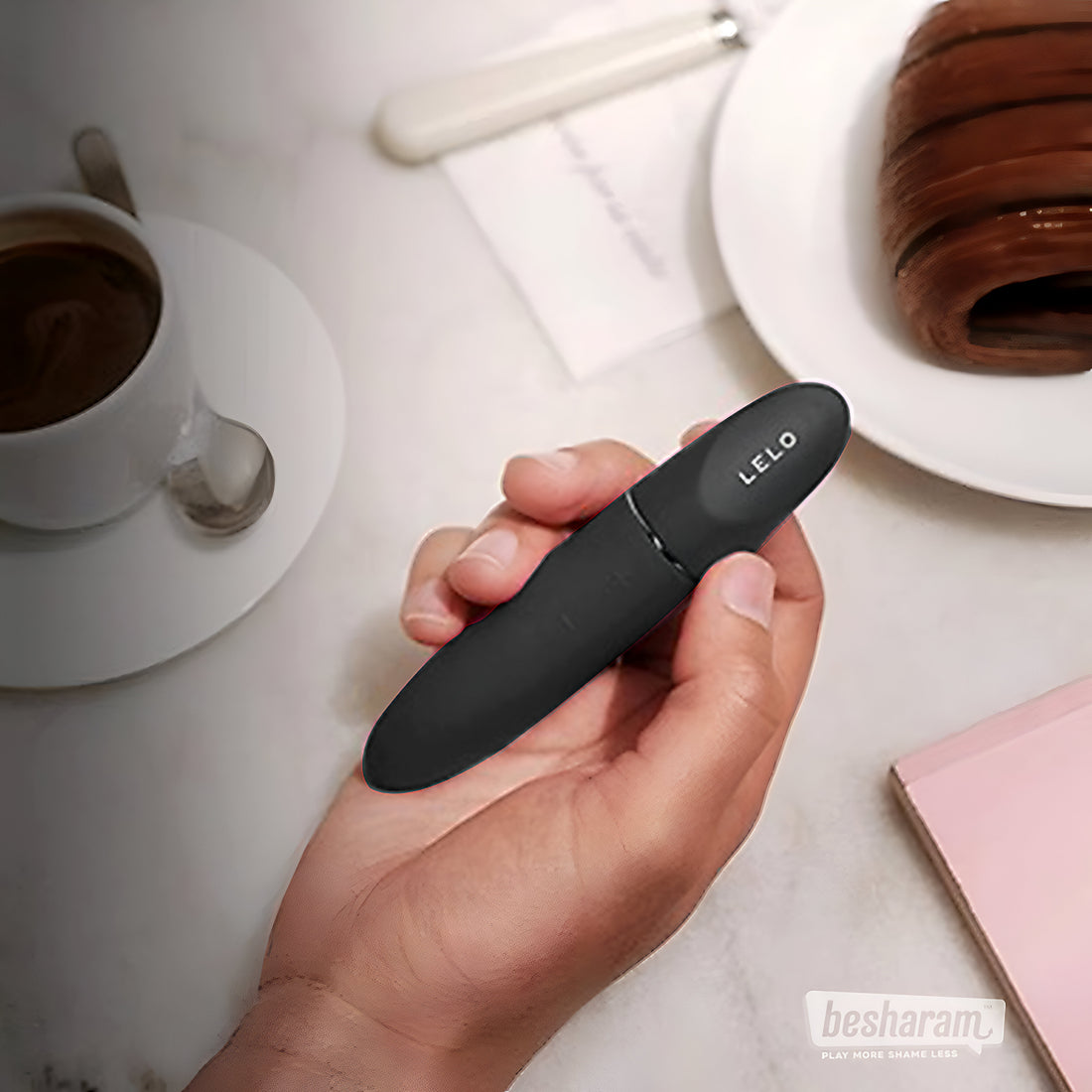 LELO Mia 3 black bullet vibrator held in hand on a coffee table with cup and notebook, sleek portable design shown in lifestyle setting