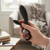 Hand holding the Satisfyer Heat Climax+ anal vibrator in a cozy living room setting, showing its sleek black dual-arm design.