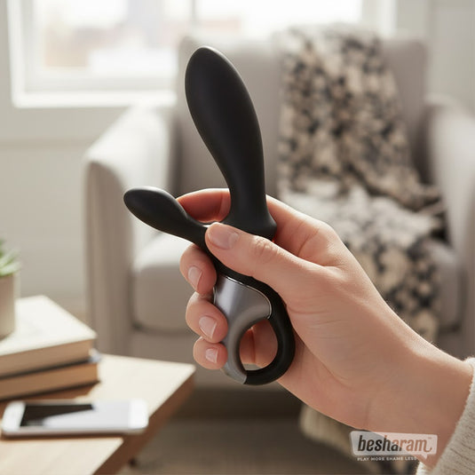 Hand holding the Satisfyer Heat Climax+ anal vibrator in a cozy living room setting, showing its sleek black dual-arm design.